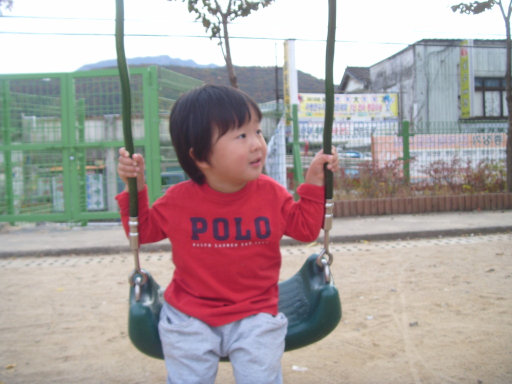 빨간 폴로 티셔츠 입은 어린 아이, 놀이터 그네에 앉아 미소.