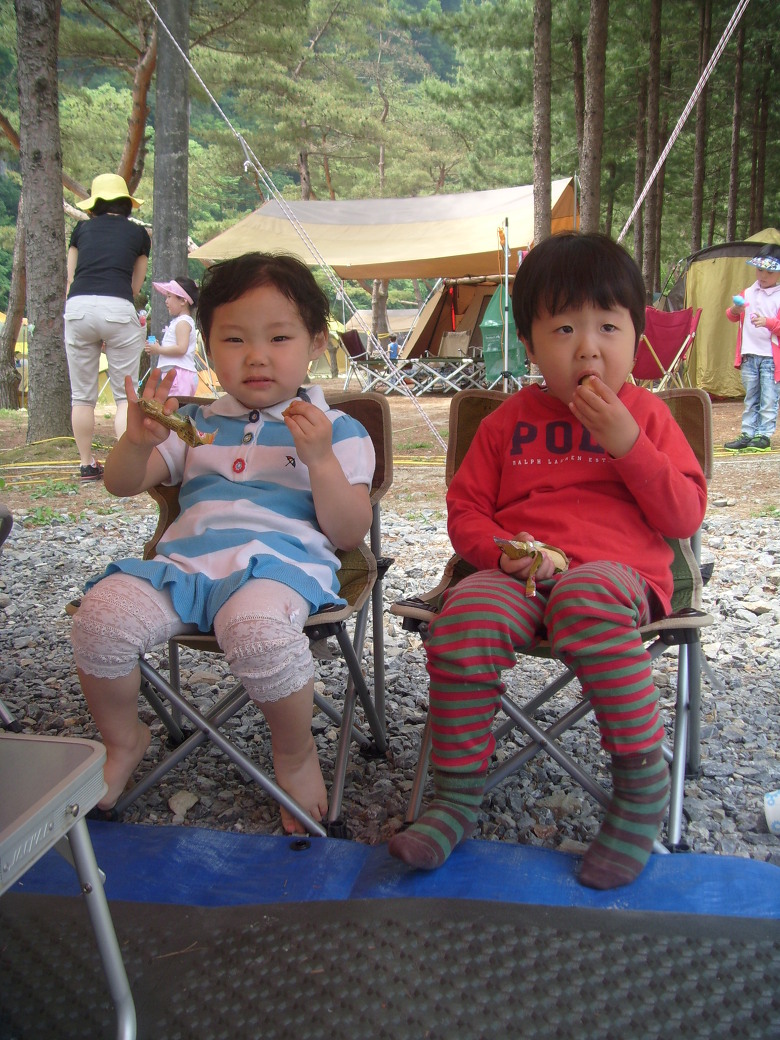 캠핑장에서 줄무늬 옷 입고 의자에 앉아 간식 먹는 두 아이들