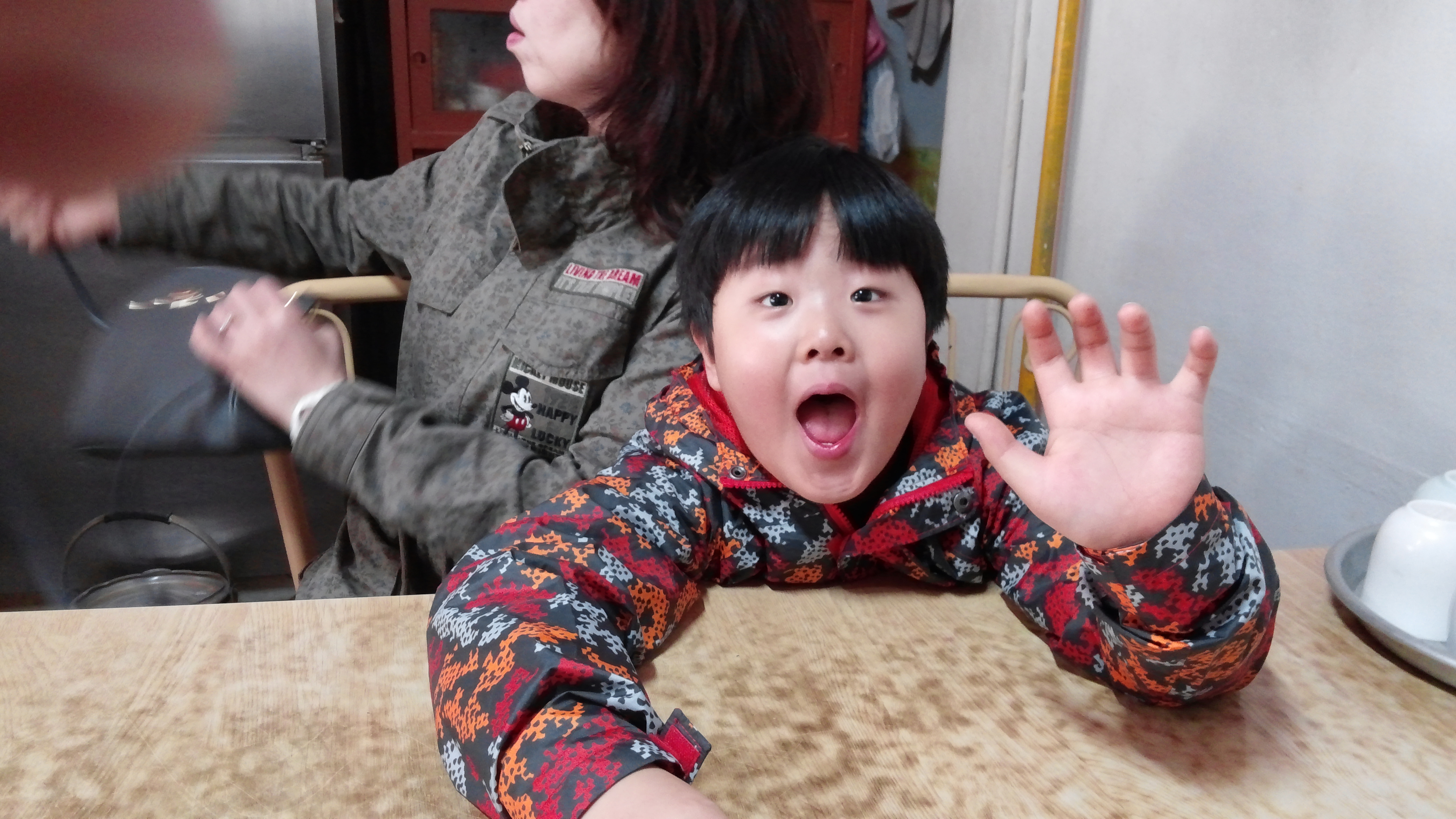 크게 입 벌린 얼굴, 손 흔드는 주황 회색 무늬 재킷, 나무 탁자