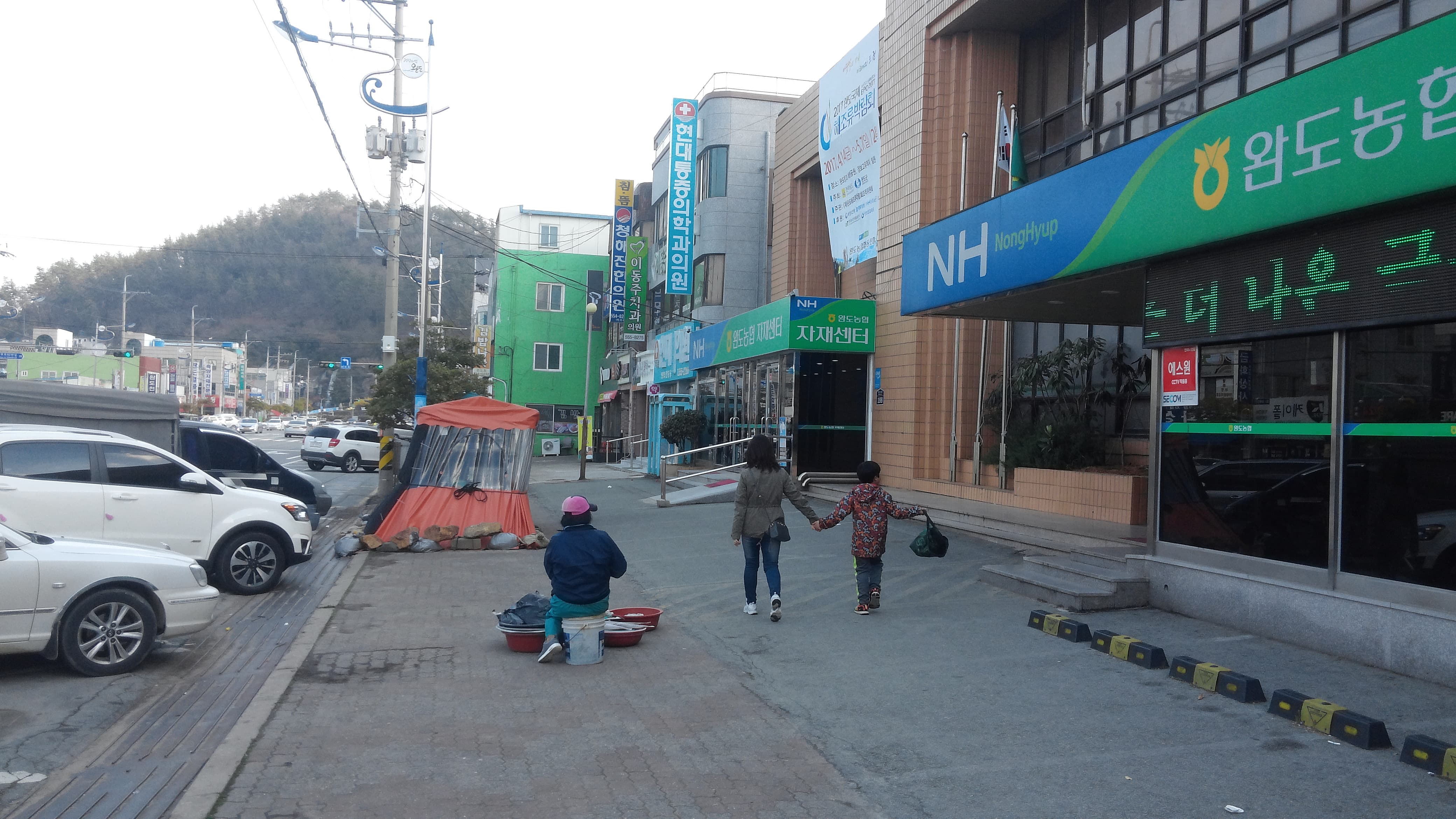 한국 도심 거리, 농협 건물, 생선 파는 상인, 두 행인, 주차 차량