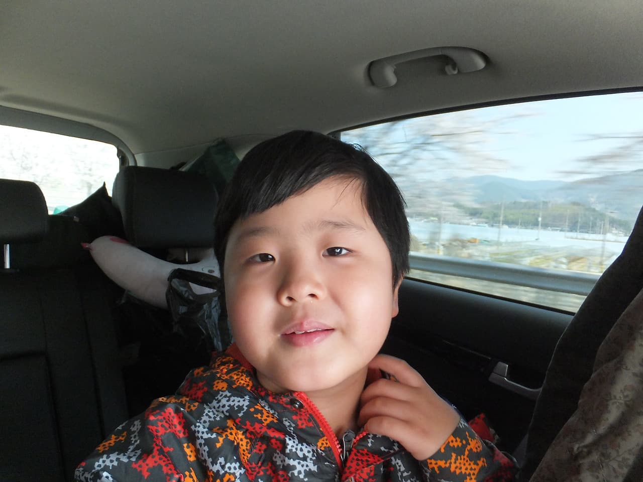밝게 웃는 아이, 차 안 컬러풀 재킷, 창밖 산과 호수