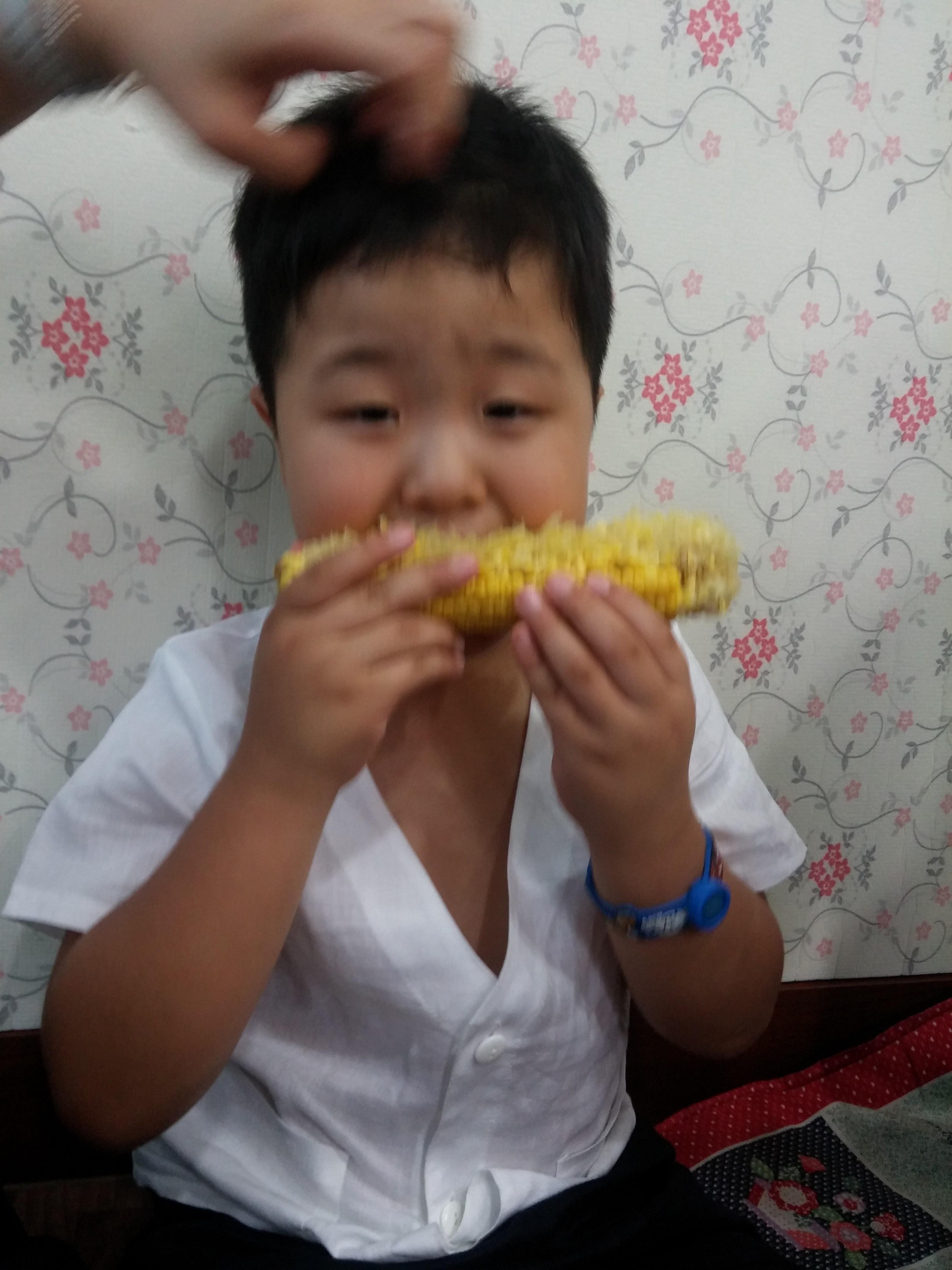 하얀 셔츠, 파란 팔찌 착용. 노란 옥수수 먹으며 머리 만지는 손.