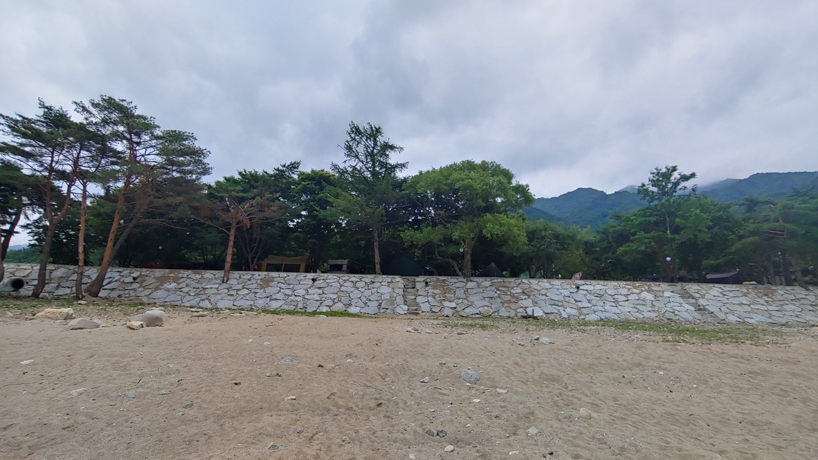모래사장, 돌담 뒤 울창한 나무와 캠핑 텐트, 흐린 산.