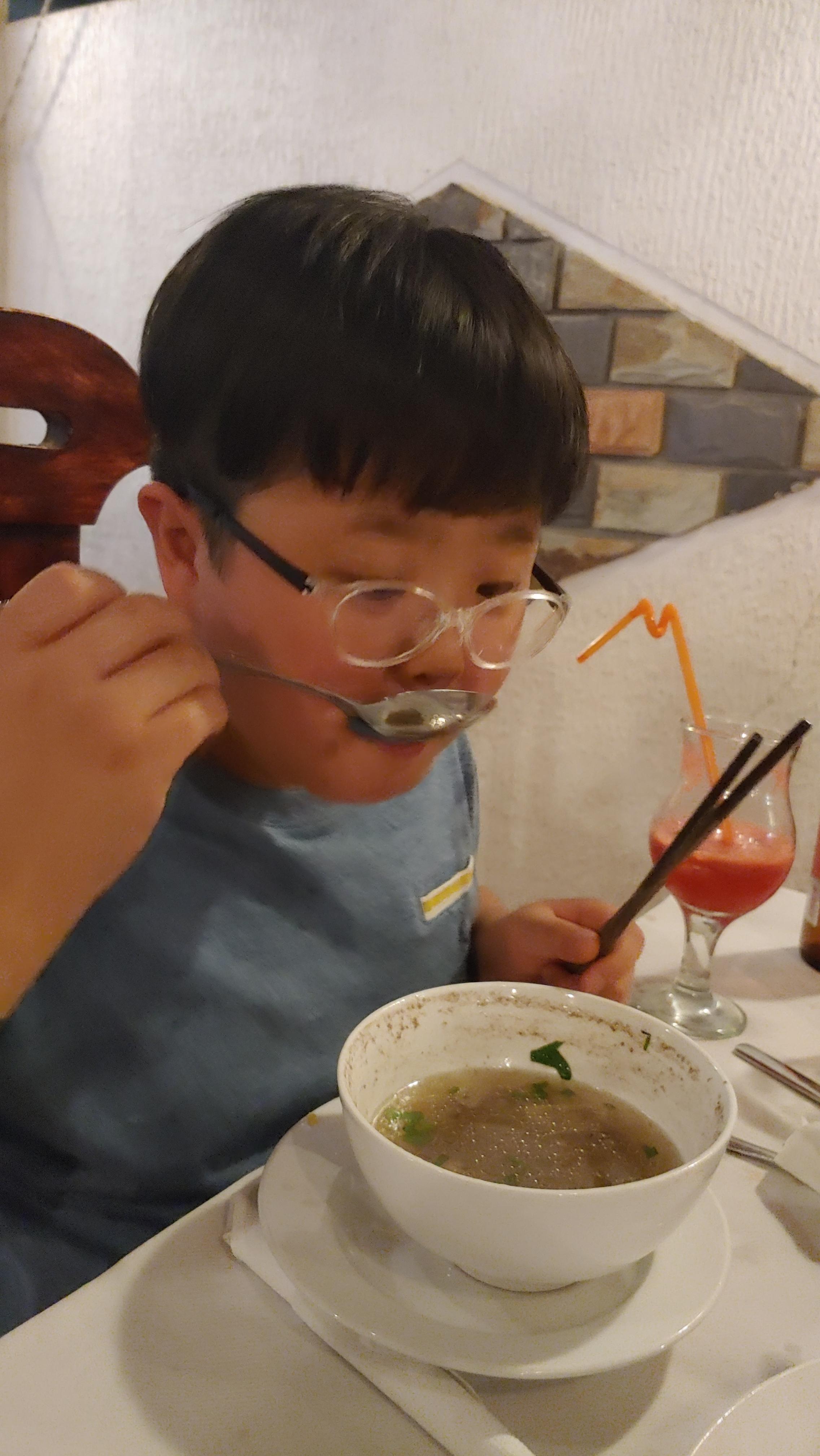 안경 쓴 이가 숟가락으로 국물 요리 먹고 젓가락 쥐고 있다.