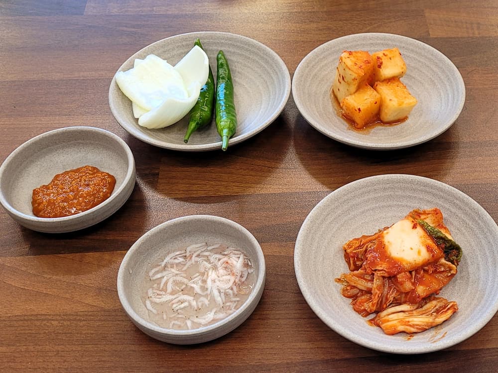 나무 식탁 위 배추김치, 깍두기, 쌈장, 새우젓, 생양파, 청고추