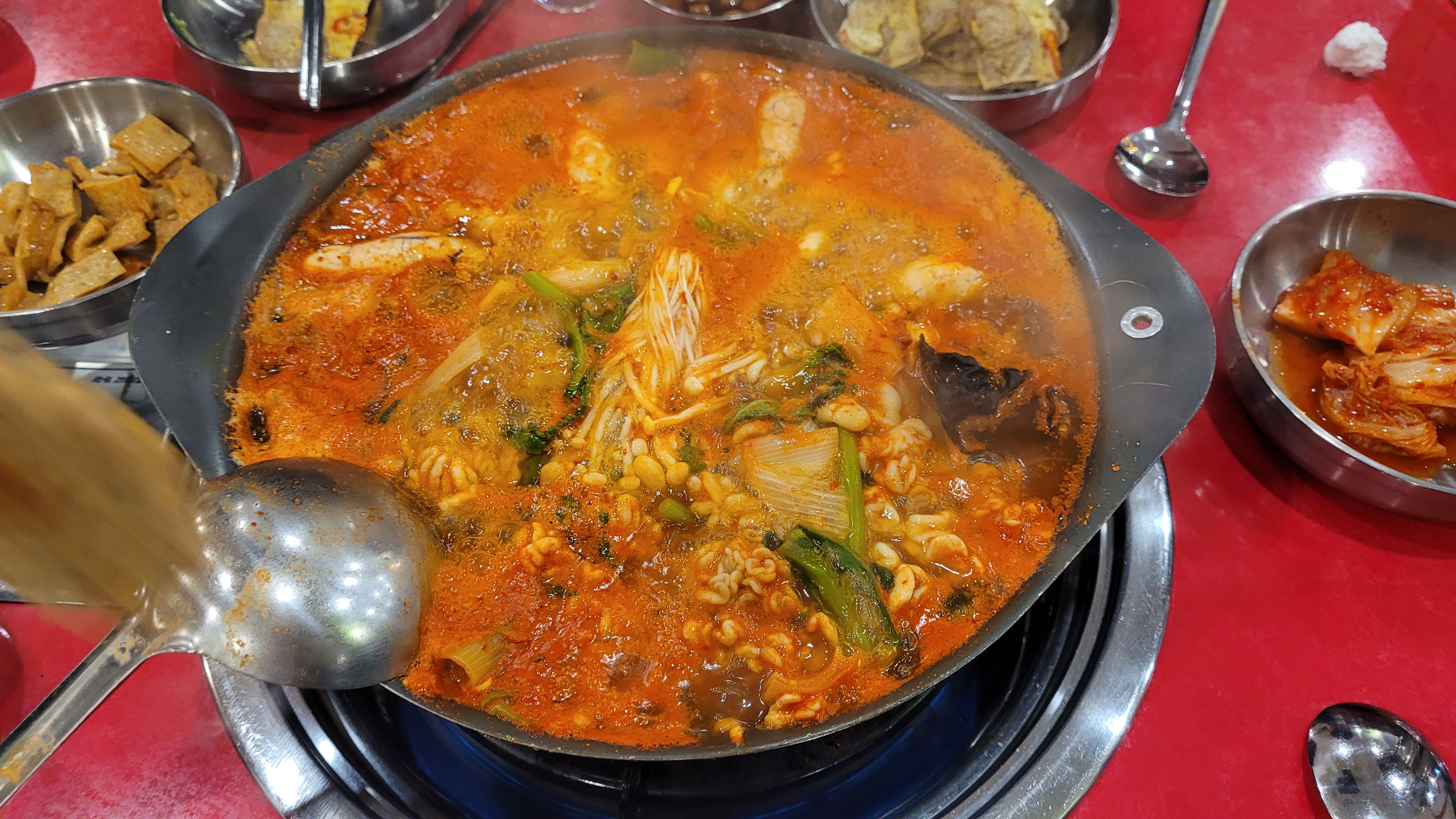 끓는 얼큰한 부대찌개, 라면, 버섯, 김치 반찬, 빨간 테이블 위
