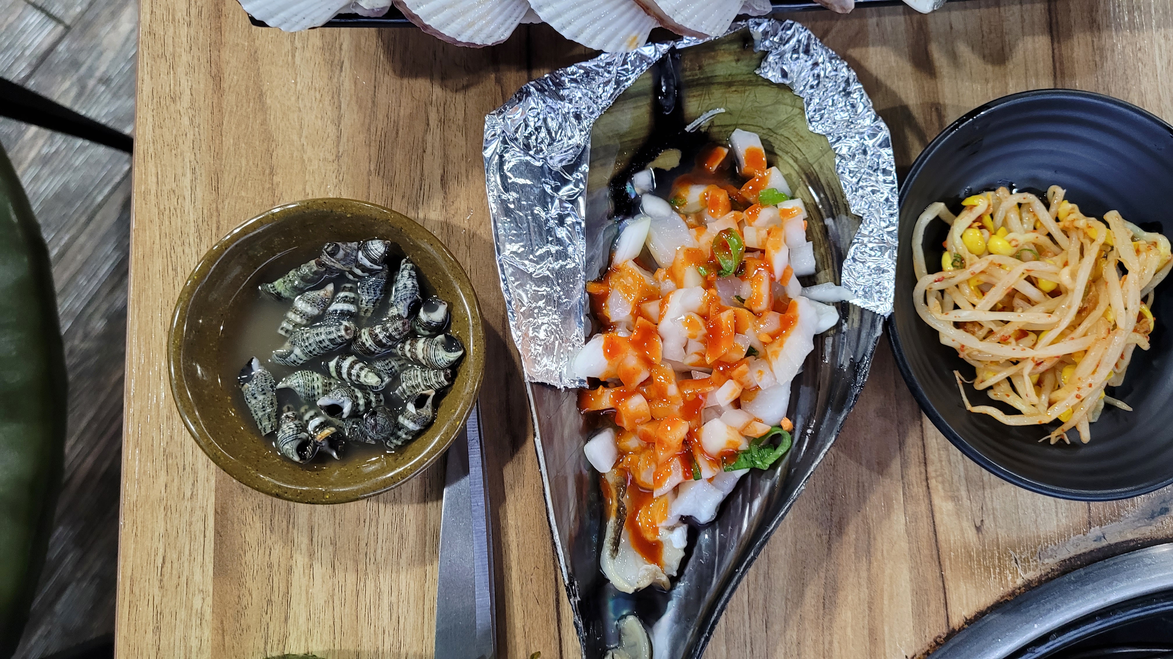 양념 조개구이, 삶은 고둥, 숙주나물, 나무 식탁