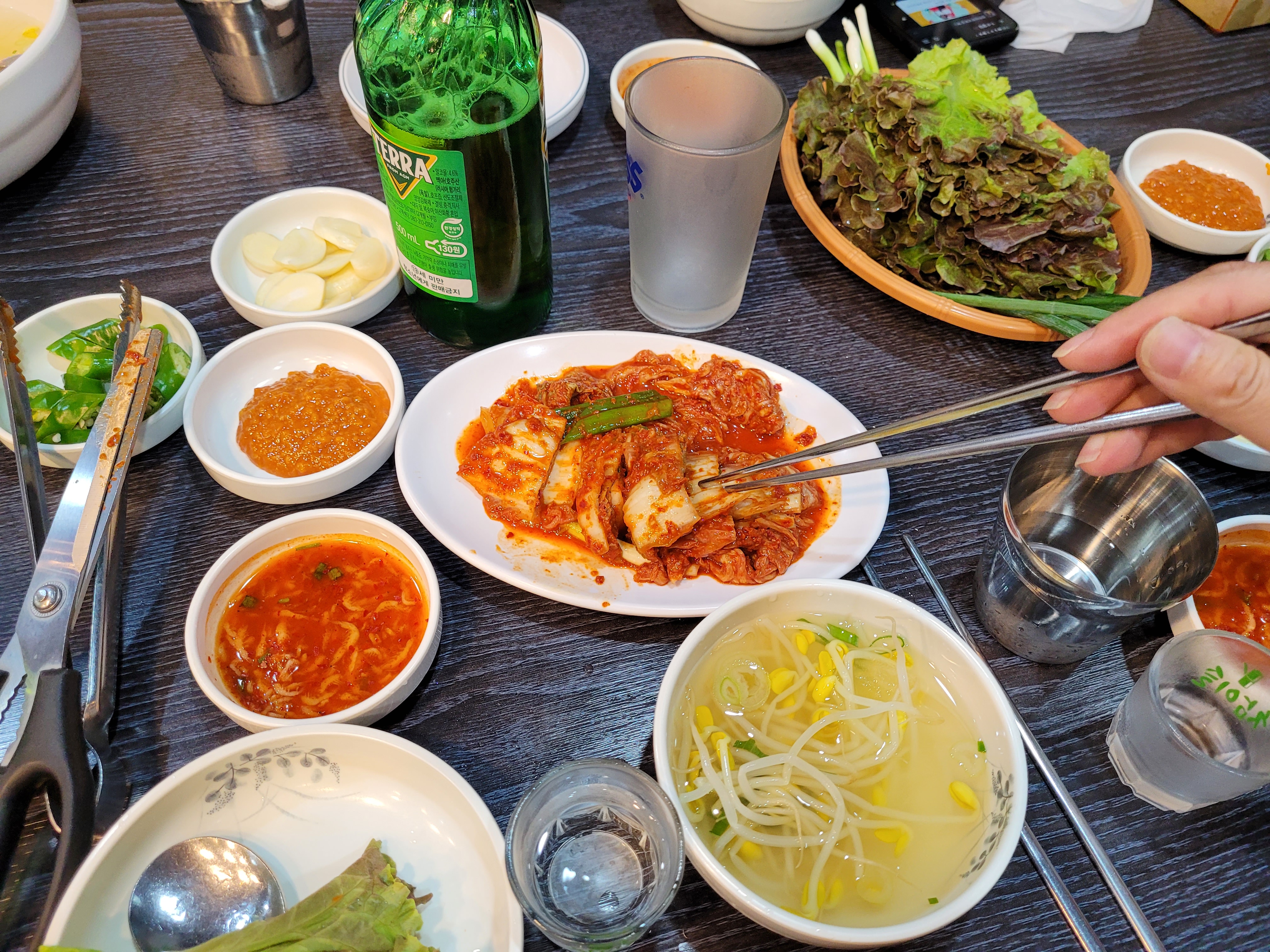 한식 식탁, 김치, 쌈채소, 콩나물국, 맥주병. 젓가락으로 김치 들기.
