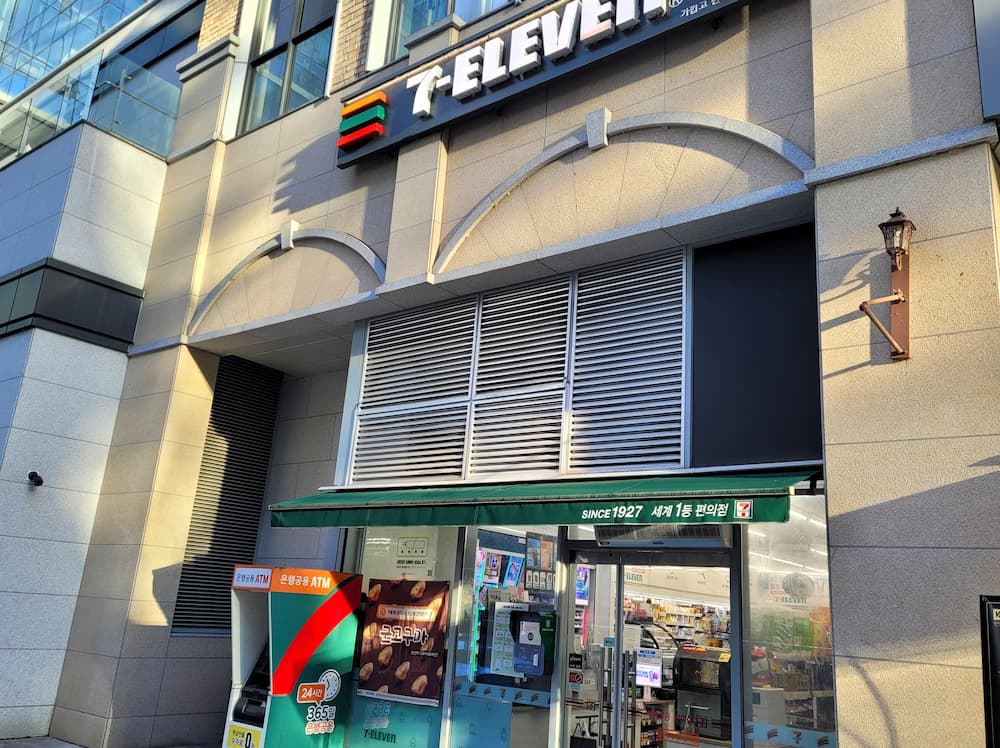 7-Eleven 편의점 입구, 녹색 차양, ATM, 현대식 건물 외관.