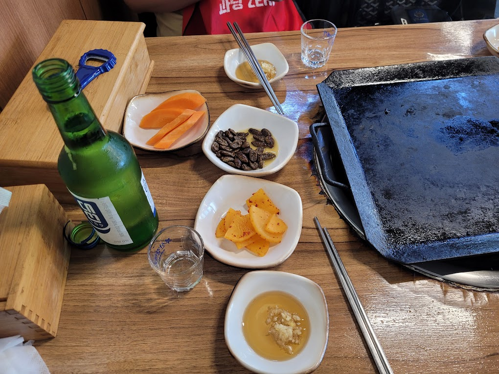 초록 소주병, 빈 고기 불판, 다양한 한식 반찬 나무 식탁