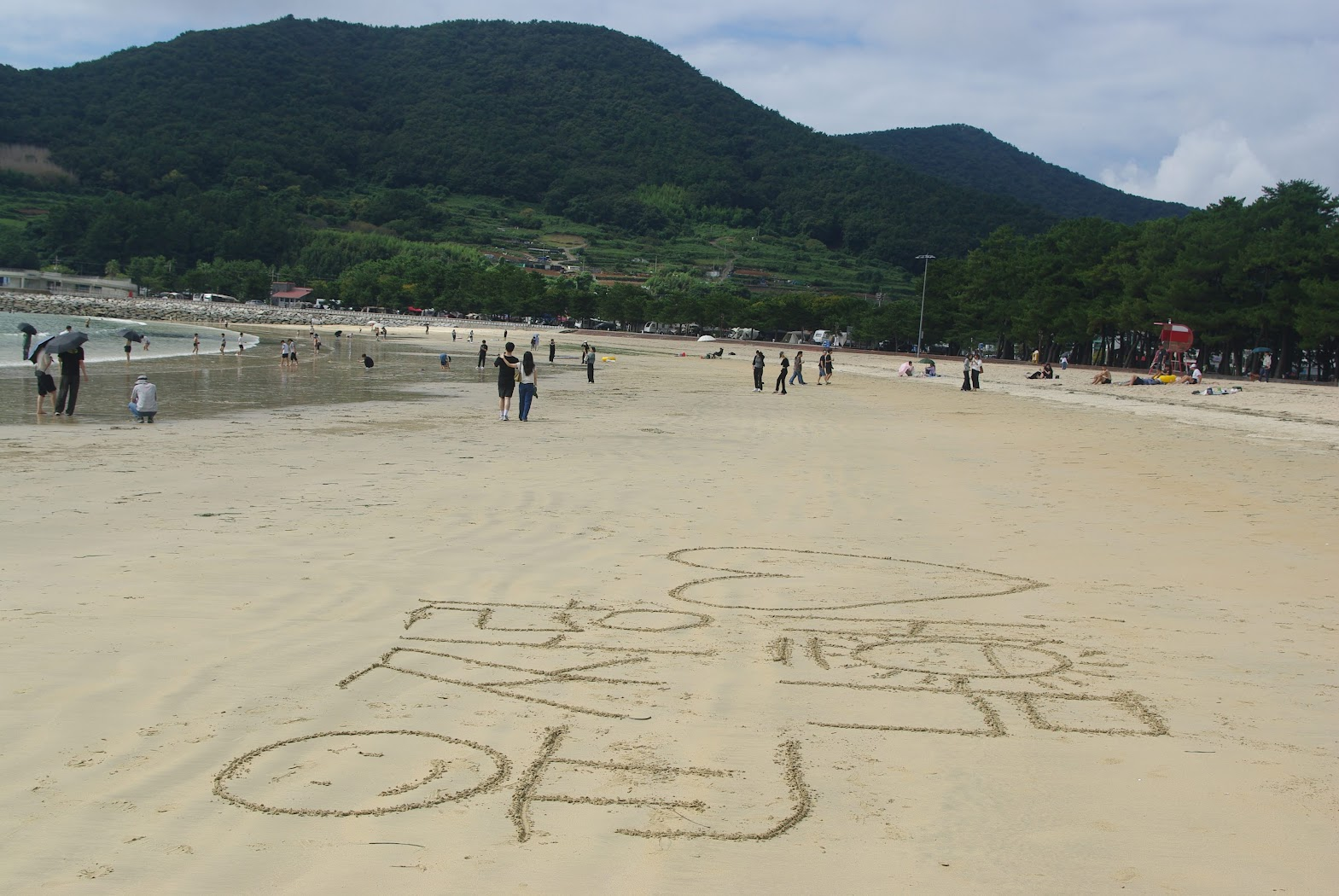 모래 해변에 "사랑해" 글씨, 하트, 미소 얼굴. 푸른 산 배경.