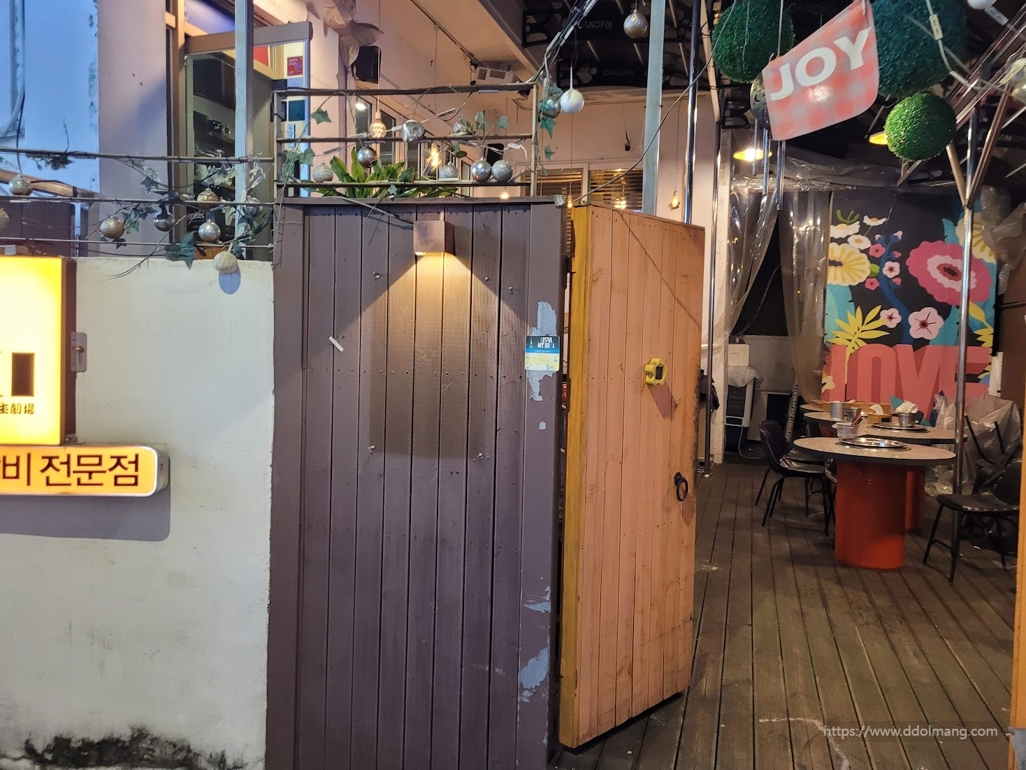 밤 식당 입구, 나무 대문, 환한 조명, 꽃 벽화, 야외 테이블