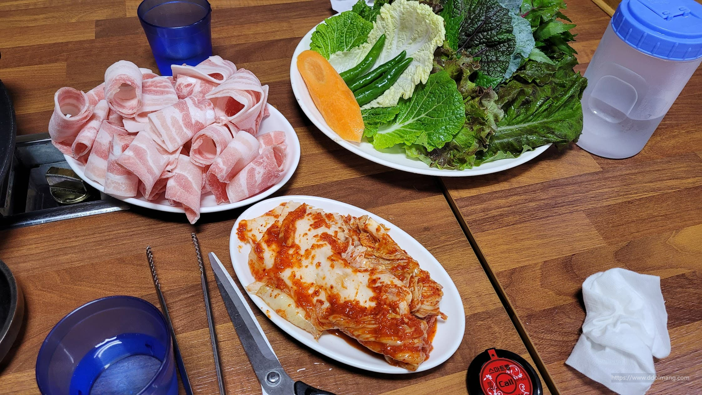 돌돌 말린 냉동 삼겹살, 쌈 채소, 김치, 나무 식탁
