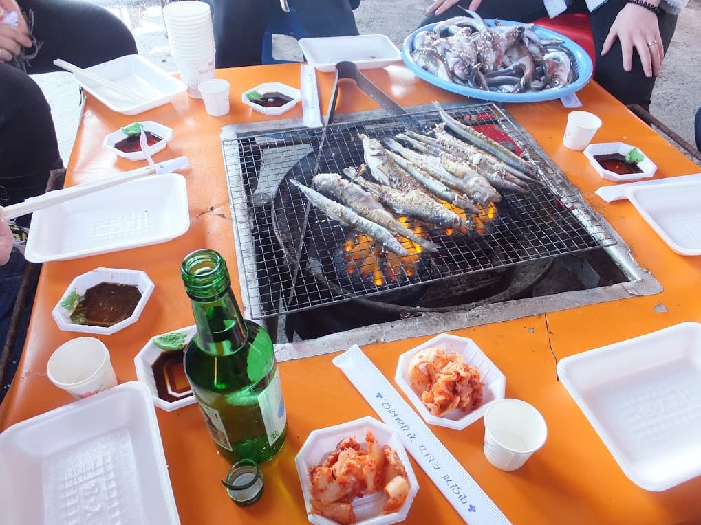 야외 식탁 위 생선 숯불구이, 날생선, 김치, 소주.