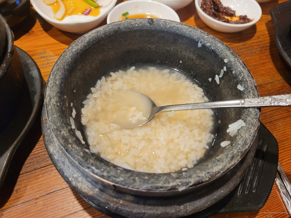 따뜻한 숭늉 담긴 검은 돌솥, 숟가락, 나무 식탁 위 반찬.
