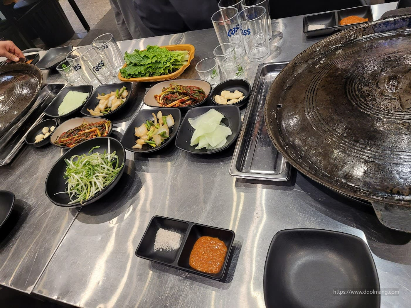 솥뚜껑 불판과 다양한 반찬, 상추, 술잔이 놓인 테이블.