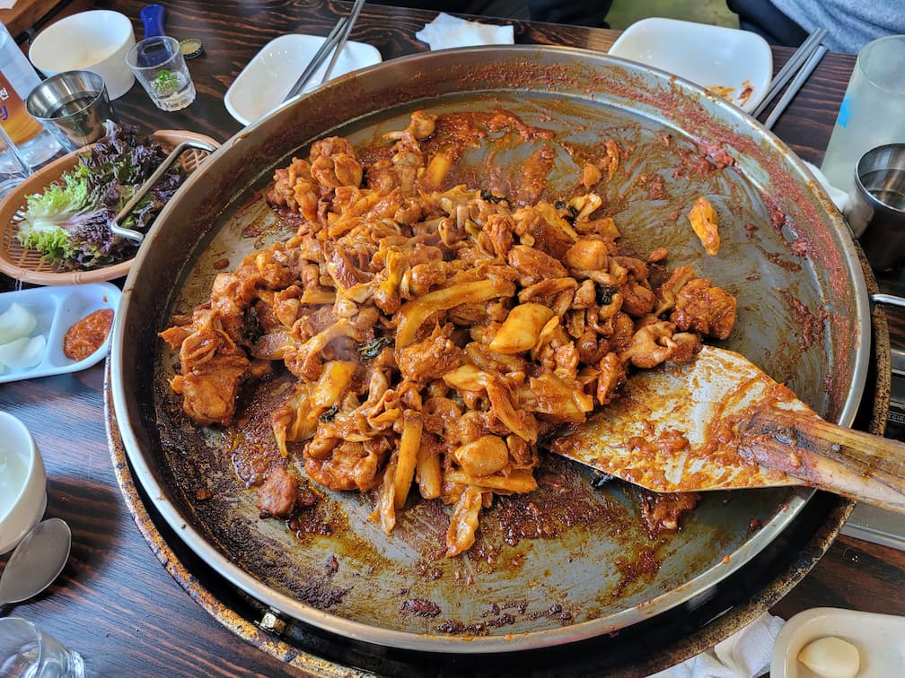 철판 매콤한 닭갈비 볶음, 주걱으로 요리 중. 신선한 쌈 채소.