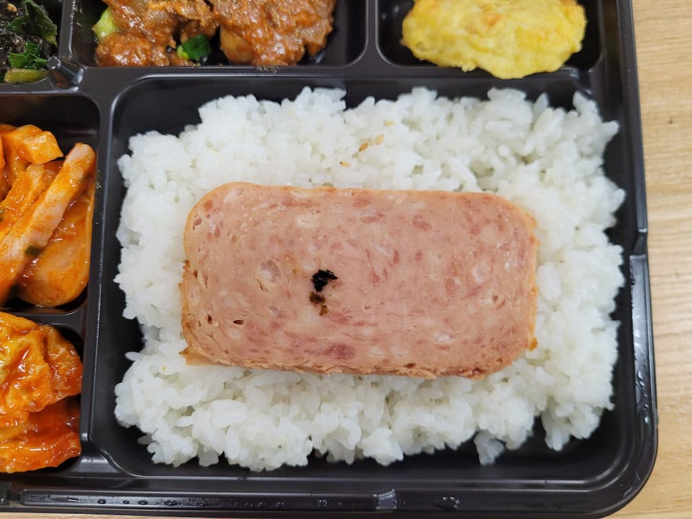 플라스틱 용기 도시락, 흰밥 위에 구운 스팸, 여러 반찬