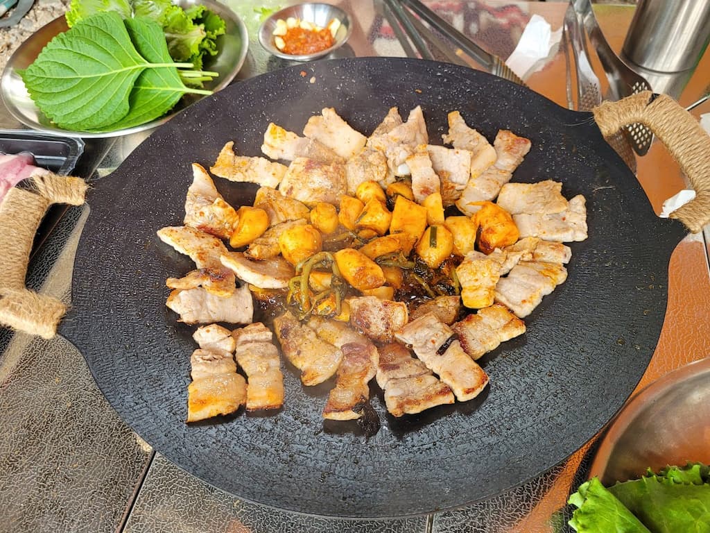 뜨거운 철판 위 노릇하게 익어가는 삼겹살, 김치, 마늘, 쌈채소