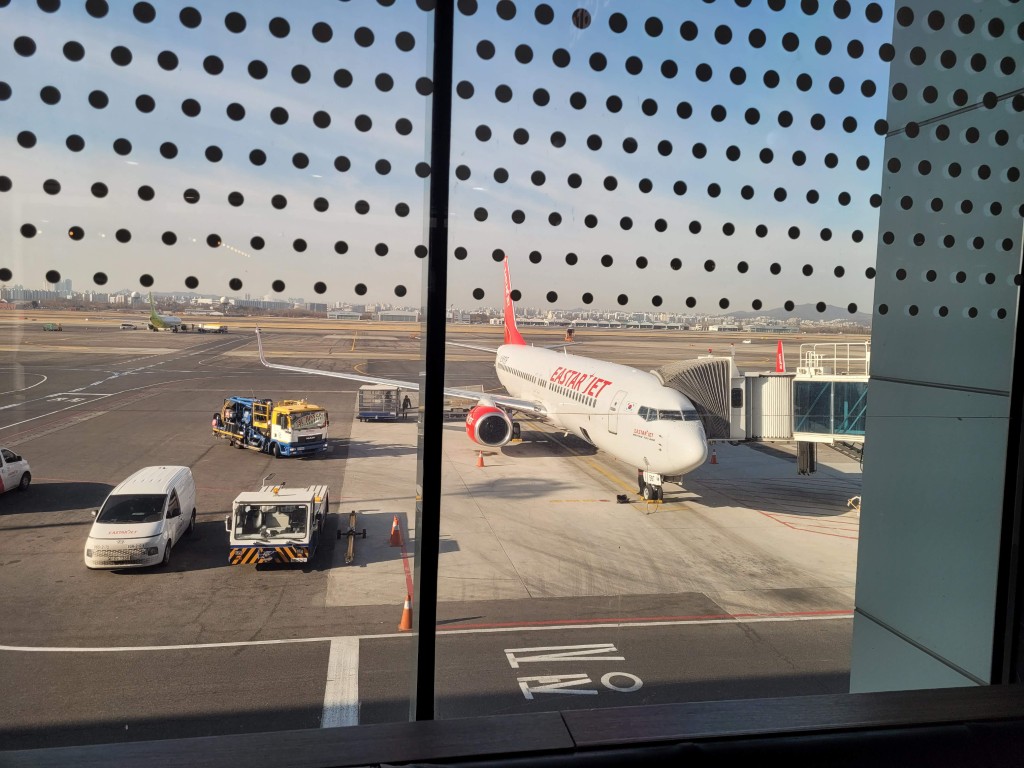 공항 터미널 창문, 이스타항공 비행기, 탑승교, 지상 차량, 맑은 하늘