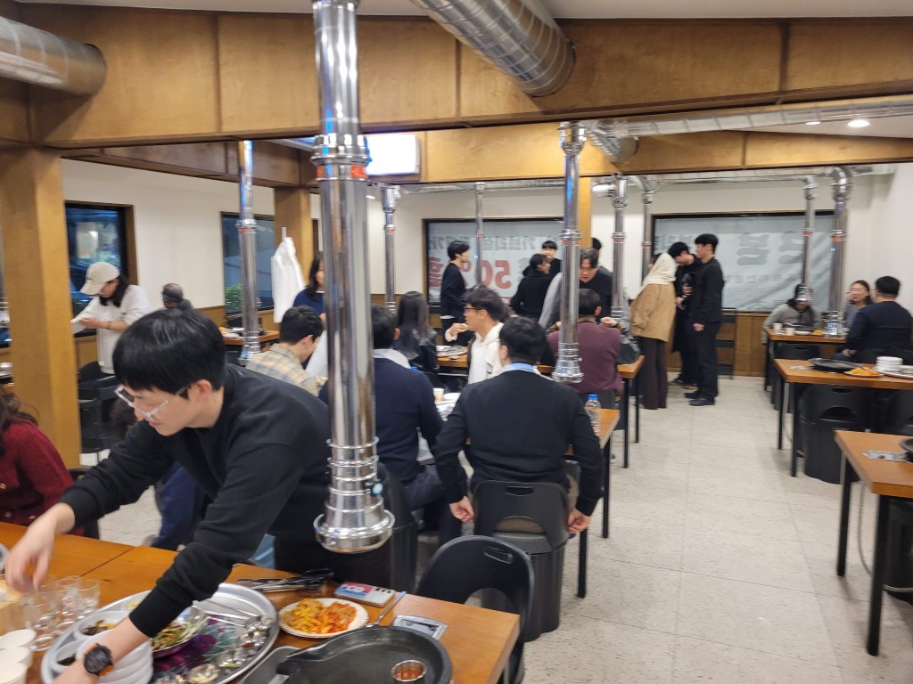 번잡한 한식 고깃집 내부, 연통 아래 식사하는 단체.