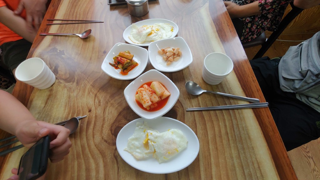 나무 식탁, 한식 반찬, 계란 프라이, 김치, 마늘, 수저.