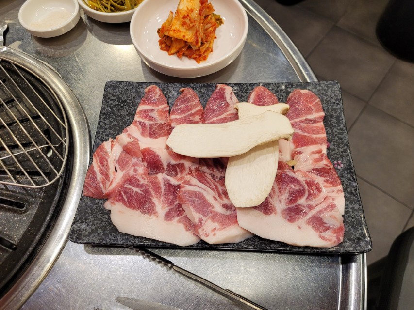 불판 옆 검은 접시 생삼겹살, 새송이버섯, 김치.