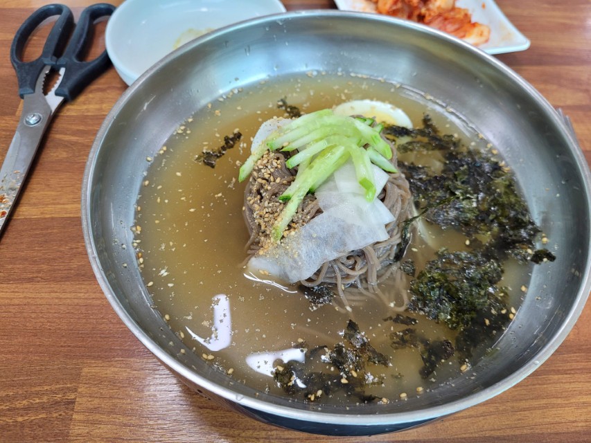 나무 테이블 위 스테인리스 그릇 시원한 물 막국수, 오이, 김가루, 가위.