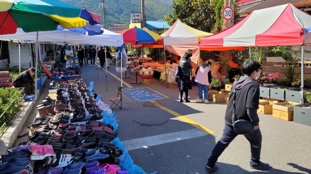 번화한 야외 시장, 다채로운 천막 아래 신발과 농산물 판매.