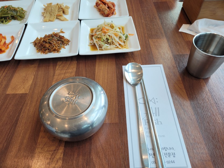 식탁 위 한국 반찬, 수저, 밥그릇 뚜껑, 물컵 정갈한 세팅.