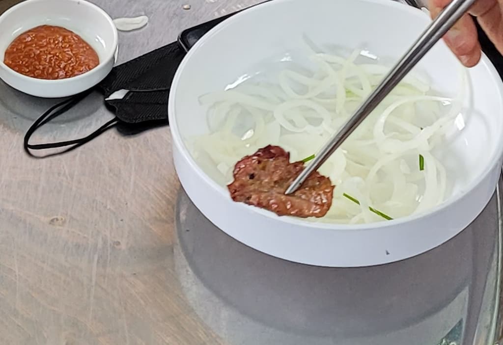젓가락 잡은 구운 고기, 얇은 양파 그릇. 옆에 매운 양념.