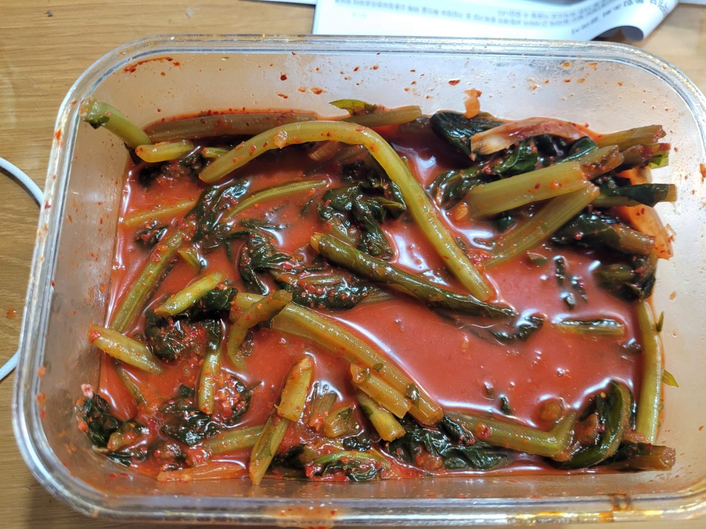 투명 용기 담긴 매콤한 붉은 총각김치, 녹색 무청