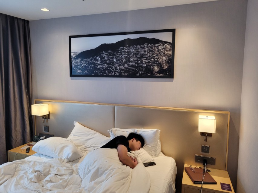 현대식 호텔 객실, 침대 위 잠든 자세, 도시 풍경 그림.