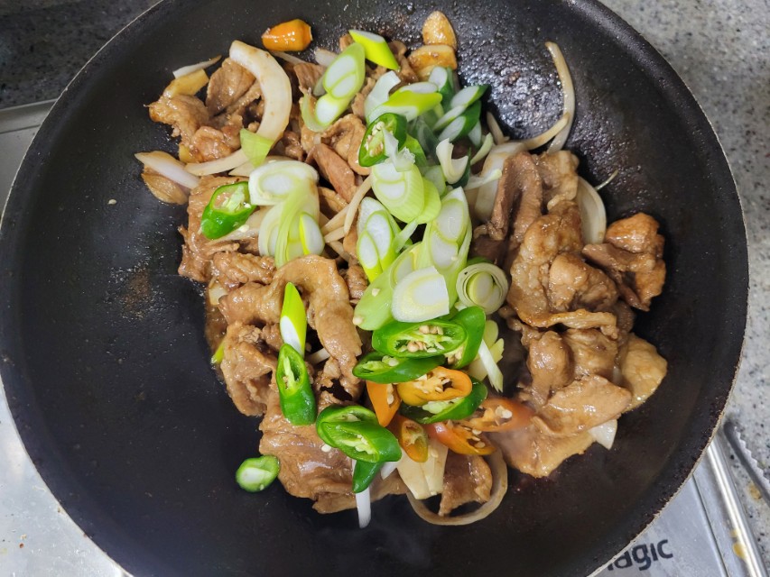 검은 팬 속 볶아지는 고기, 양파, 대파, 풋고추