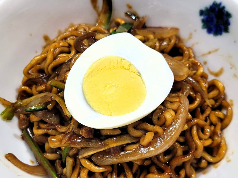 흰 그릇 짜장면, 검은 소스 면, 반쪽 삶은 계란, 양파 오이