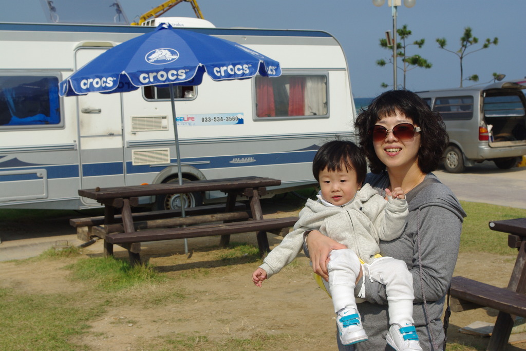 선글라스 엄마 아기 캠핑카 앞 크록스 파라솔 아래 미소.
