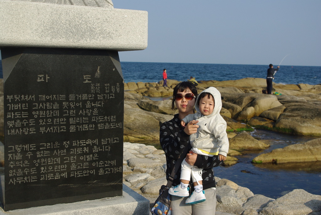 바위 해안 비석 옆 아기 안은 보호자, 푸른 바다