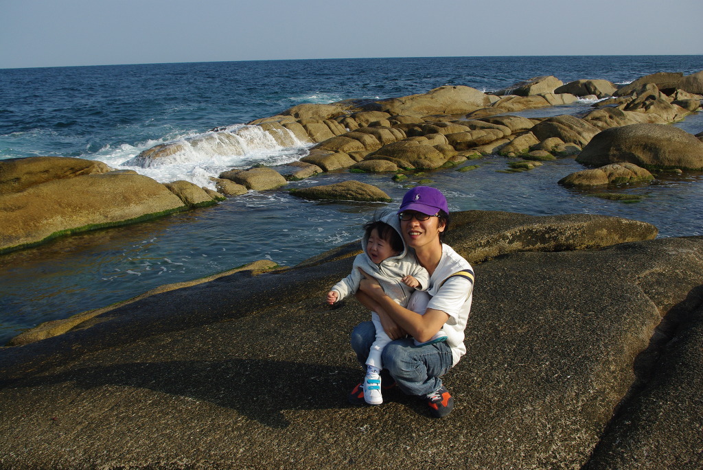 바다 바위 해변, 보라색 모자 쓴 이가 유아 안고 행복한 미소.