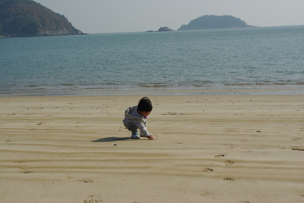 모래 해변 웅크린 유아, 막대기로 모래에 그림, 바다 배경.