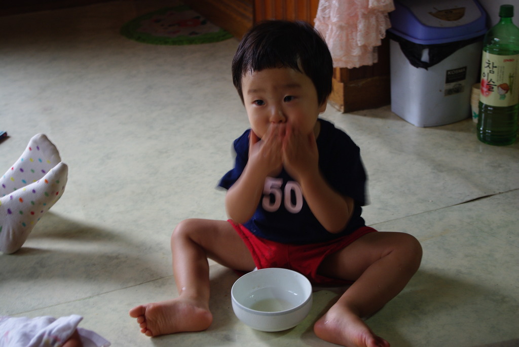 바닥에 앉은 아이, 파란 티셔츠 입고 앞에 그릇 둔 채 입 가림