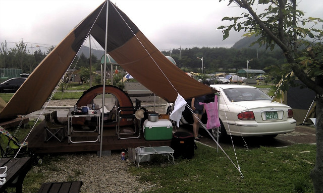 야외 캠핑장 갈색 타프, 텐트, 흰색 차량, 캠핑 장비